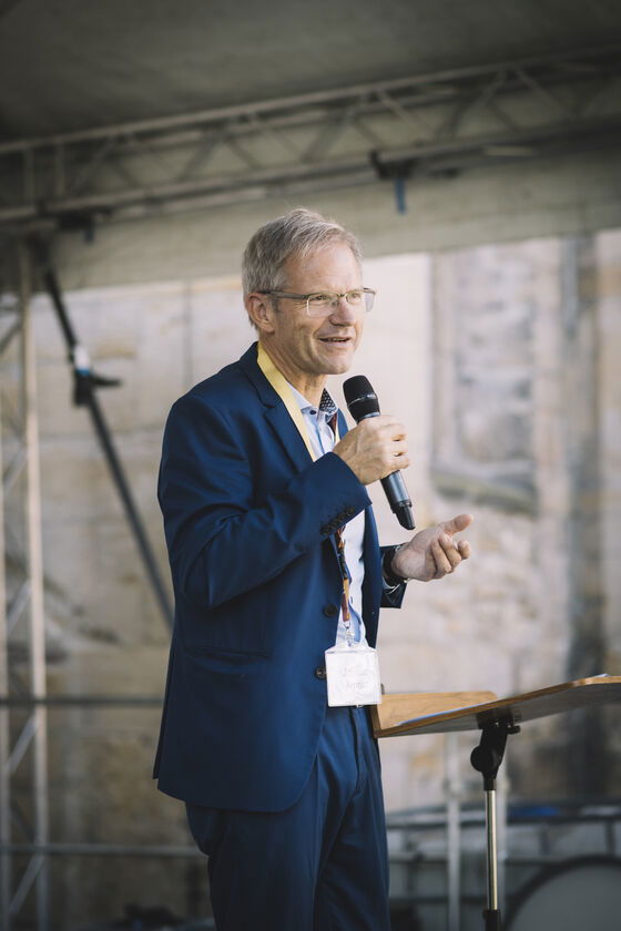 Eine als Mann lesbare Person im Anzug spricht in ein Mikrofon.