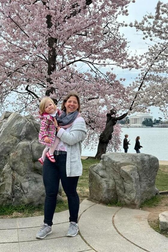 Eine weiblich gelesene Person mit kleinem Kind auf dem Arm steht vor rosa blühenden Bäumen.