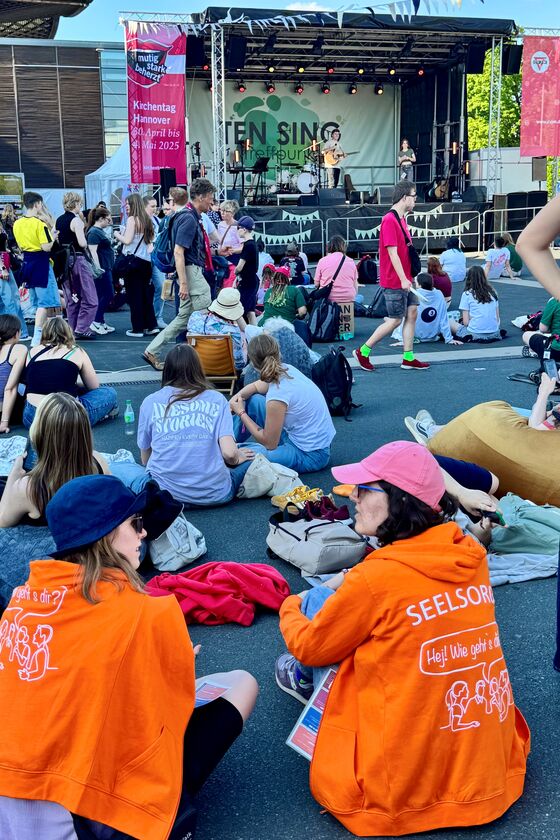 Personen sitzen und stehen vor einer Bühne, auf der eine männlich zu lesende Person Gitarre spielt.