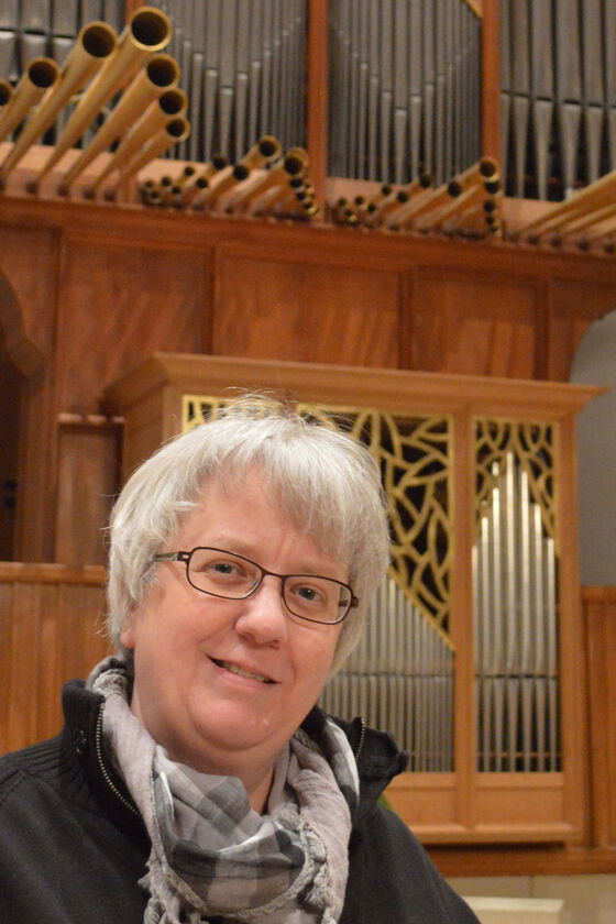 Eine weiblich gelesene Person mit grauem Haar und Halstuch vor einer Orgel.