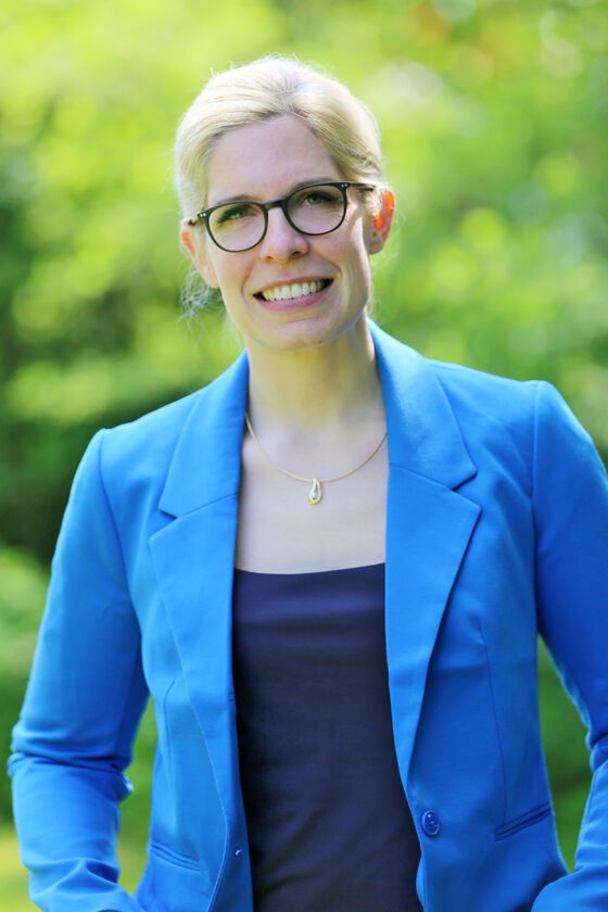 Eine weiblich gelesene Person mit blonden Haaren und Brille in hellblauem Blazer und dunklem Top.