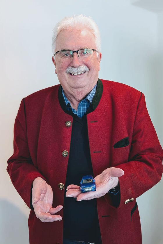 Eine männlich gelesene Person mit weißen Haaren und Schnauzer in roter Jacke, die ein kleines Auto in Händen hält.