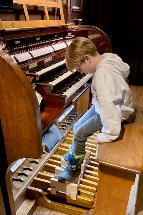 Ein Kind sitzt vor einer Orgel-Tastatur und erreicht mit den Füßen geradeso die Pedale.