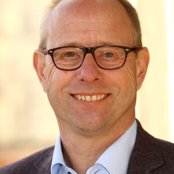 Ein Mann mit Brille mit dunkler Fassung, hoher Stirn und kurzen Haaren. Er trägt ein hellblaues Hemd und ein Jackett.