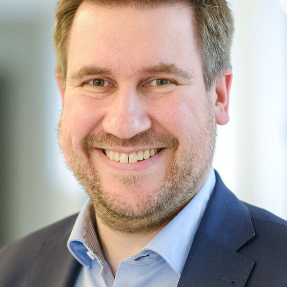 Das Bild zeigt eine männlich lesbare Person mit kurzen, mittelbraunen Haaren im Portrait. Die Person trägt einen Bart, ein hellblaues Hemd und ein dunkelblaues Sakko.