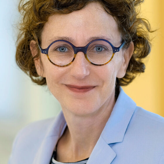 Das Bild zeigt eine weiblich lesbare Person mit dunklen kurzen lockigen Haaren, einer Brille mit dunkler Fassung, einem blau-weiß geringeltem Oberteil und einem hellblauen Jackett darüber. 