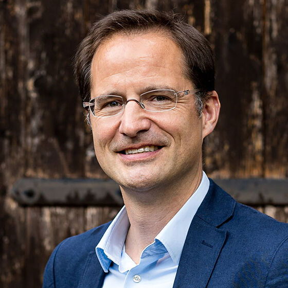 Eine männlich lesbare Person mit Brille, blauem Hemd und Sakko ist im Portrait zu sehen.