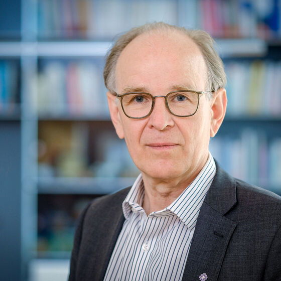 Eine männlich lesbare Person ohne Bart mit grauen Haaren und hohem Haaransatz, einer Brille mit brauner Fassung, einem hellen gestreiften Hemd und einem dunklen Sakko; im Hintergrund eine Bücherwand.