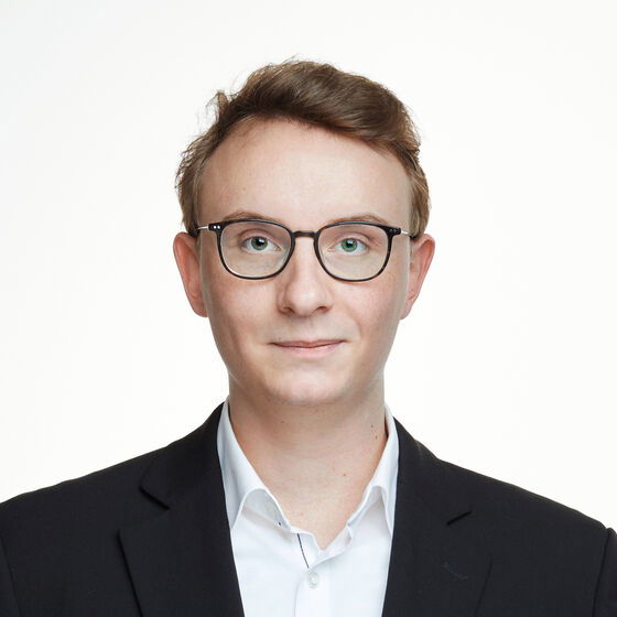 Ein weißer, junger Mann mit Brille lächelt verhalten. Er trägt ein dunkles Sakko und ein weißes Hemd.