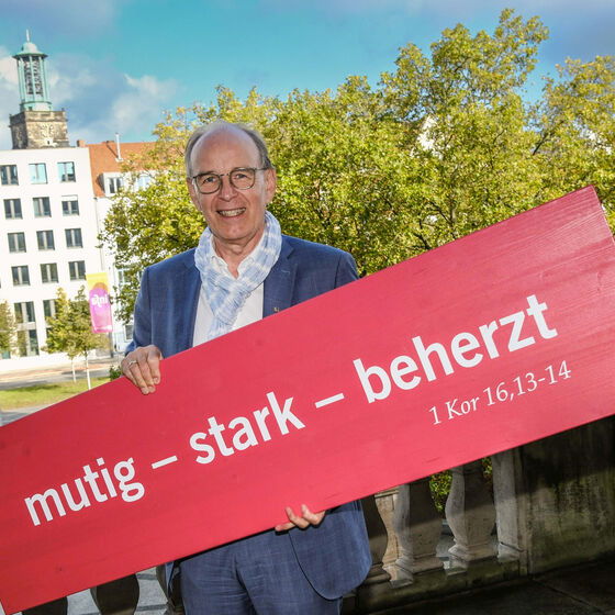 Eine als Mann lesbare Person hält ein Plakat mit der Kirchentagslosung "mutig - stark - beherzt"