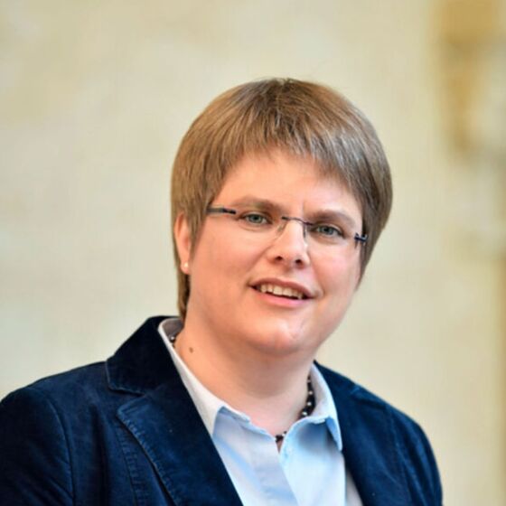 Eine weiblich gelesene Person mit kurzen Haaren und Brille in hellblauer Bluse und dunkelblauem Blazer schaut freundlich in die Kamera.