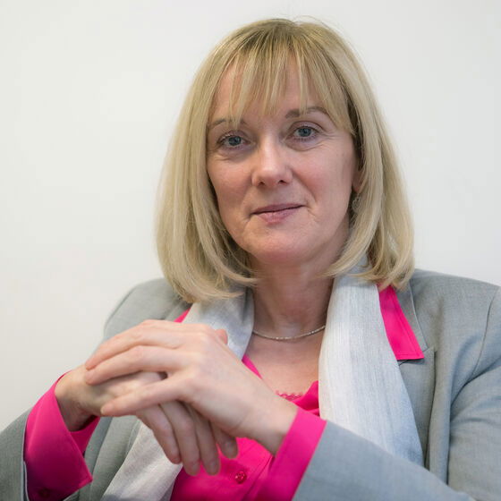 Eine als Frau lesbare Person mit Pagenfrisur sitzt mit übereinanderliegenden Händen.