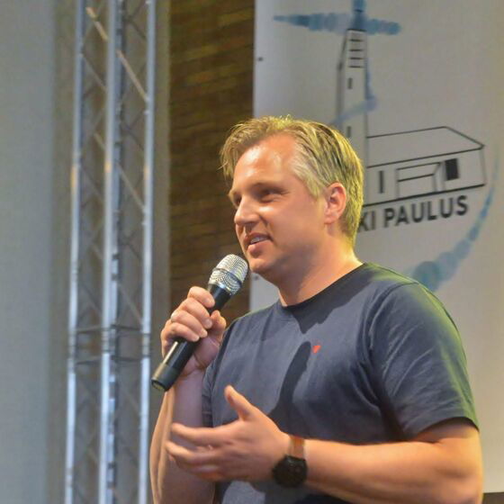 Eine männlich gelesene Person mit kurzen blonden Haaren hält ein Mikro in der Hand und spricht.