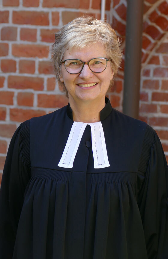 Eine weiblich gelesene Person mit kurzen blonden Haaren im Talar steht vor einer Backsteinwand, vermutlich eine Kirche.