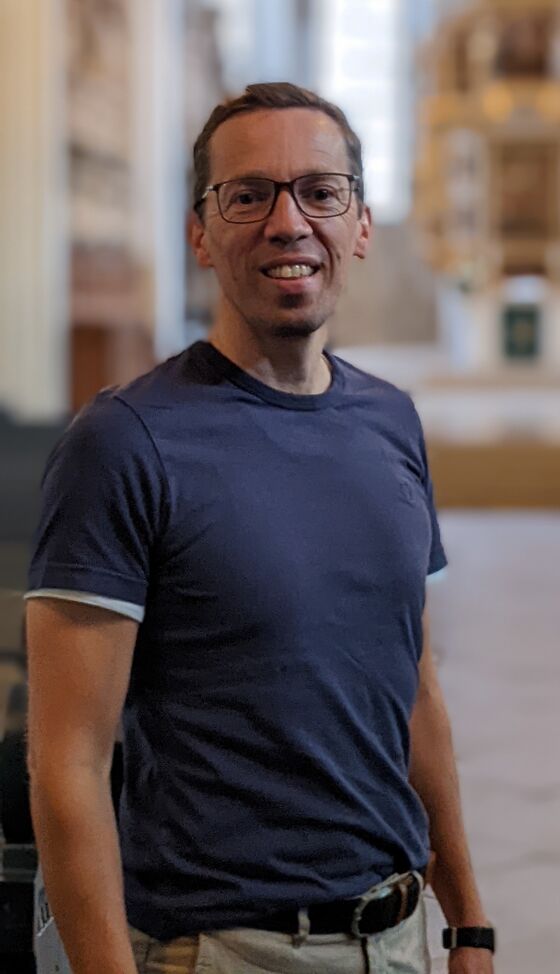 Eine männlich gelesene Person in blauem T-Shirt und mit Brille steht in einem Mittelgang einer Kirche.
