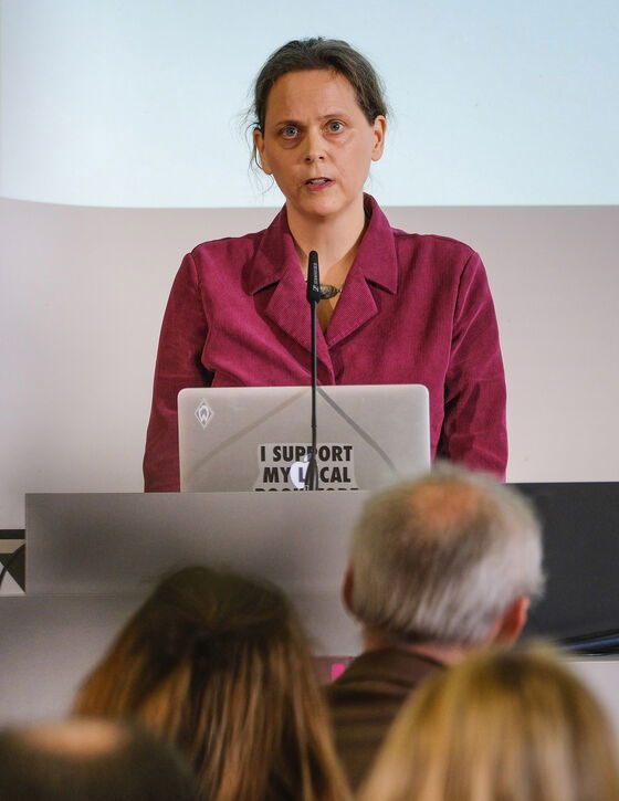 Eine als Frau lesbare Person steht an einem Redepult und spricht.