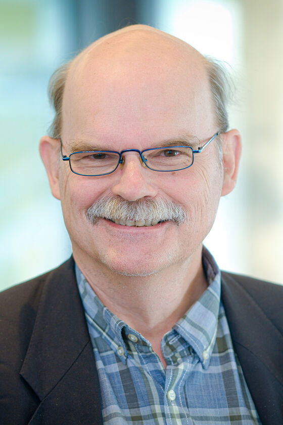 Eine männlich gelesene Person mit Brille, Schnauzbart und blau-kariertem Hemd lächelt.