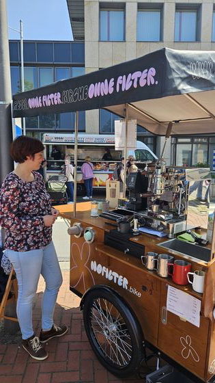 Eine weiblich gelesene Person steht neben einer mobilen Kaffee-Bar mit Siebträgermaschine und einem schwarzen Dach auf dem steht: Reden ohne Filter, Kirche ohne Filter.