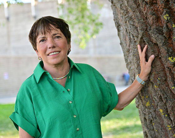 Eine als Frau lesbare Person in grüner Bluse lehnt an einem Baum.
