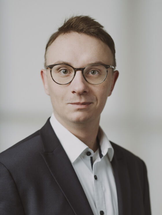 Eine als Mann lesbare Person im Jackett, weißen Hemd und mit Brille