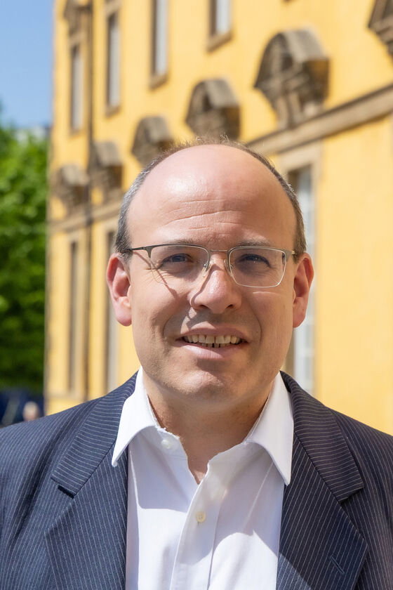 Eine als Mann lesbare Person mit hoher Stirn und Brille steht vor einem gelben Haus.