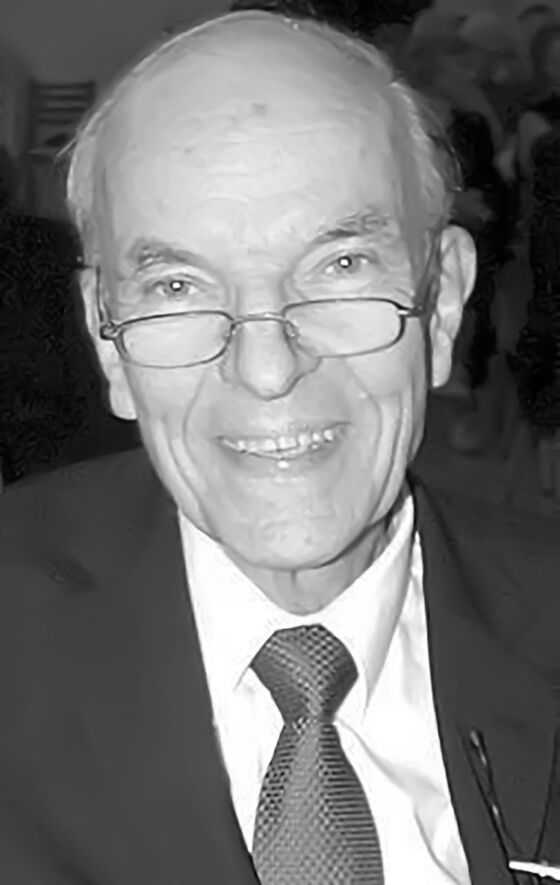 Eine männlich lesbare Person ohne Bart, mit Brille und Halbglatze, in hellem Hemd und schwarzem Sakko und mit schwarzer Krawatte (sw-Bild).
