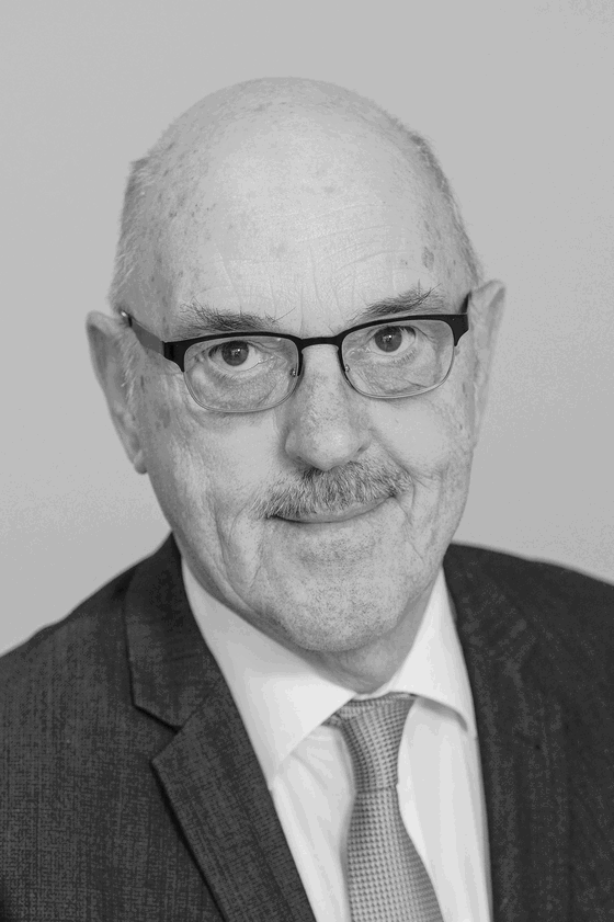 Das Schwarzweiß-Bild zeigt eine männlich lesbare Person mit Schnurrbart, wenig Haaren und einer Brille mit dunkler Fassung. Die Person trägt ein weißes Hemd, eine graue Krawatte und ein dunkles Sakko.