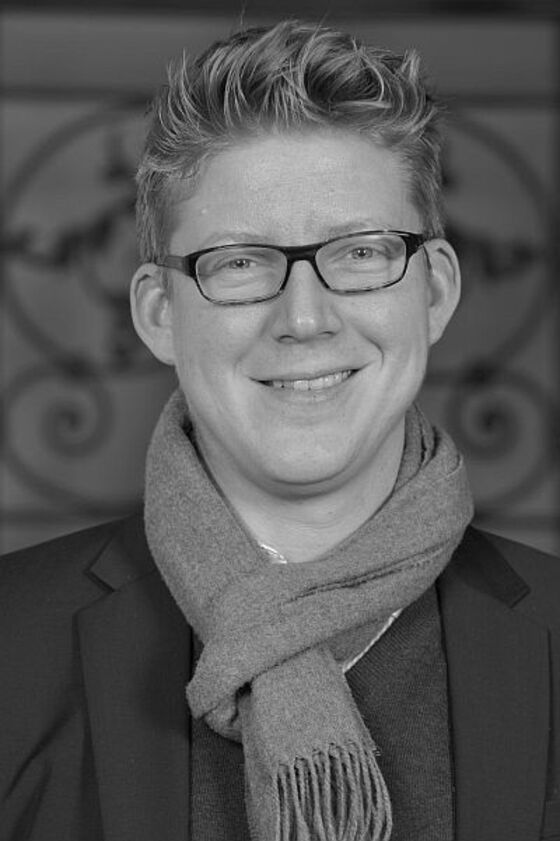 Das Schwarzweiß-Bild zeigt eine männlich lesbare Person mit lockigen kurzen Haaren und einer Brille mit dunkler Fassung. Die Person trägt einen Schal um den Hals und ein dunkles Oberteil.