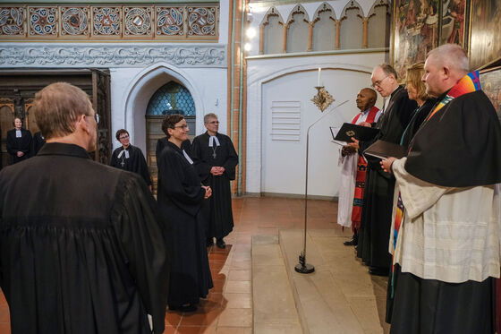 Ein weiblich zu lesende Person im Talar steht vor dem Altar. Auf der Altarstufe stehen mehrere männlich zu lesende Personen in geistlichen Gewändern.