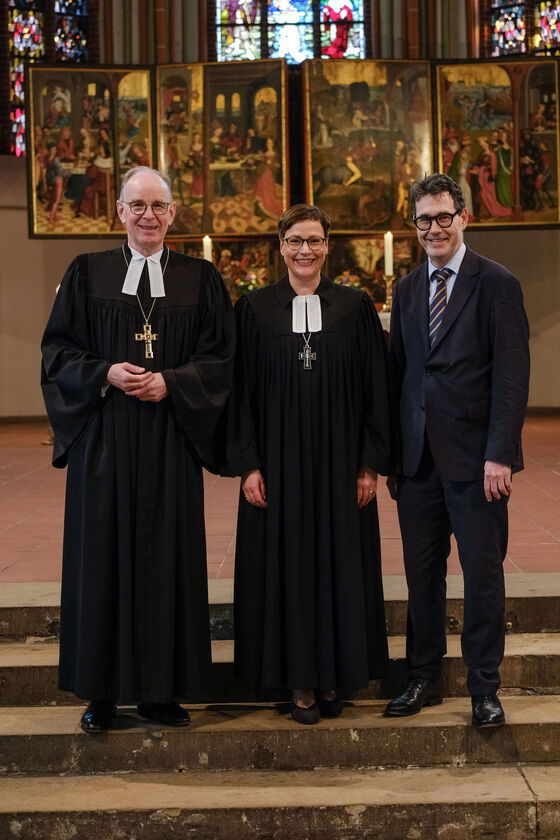 Zwei männlich zu lesende Personen und eine weiblich zu lesende Person stehen auf Treppenstufen. Im HIntergrund ist ein Altar zu sehen.
