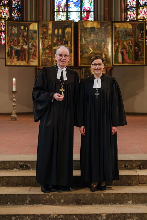 Eine männlich und eine weiblich zu lesende Person im Talar stehen vor einem Altar.