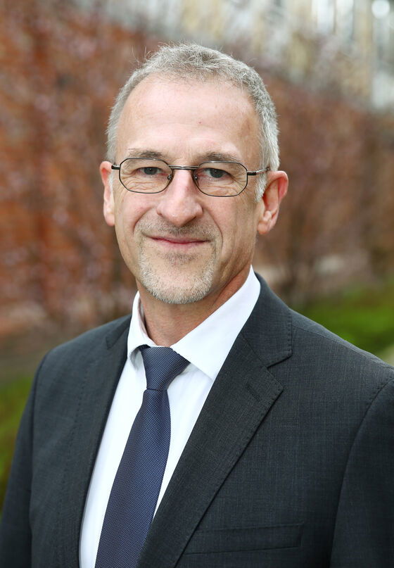 Eine männlich gelesene Person mit kurzen grauen Haaren, grauem kurzen Vollbart und Brille in einem schwarzen Sakko und mit weißem Hemd. 