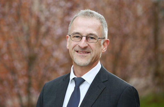 Eine männlich gelesene Person mit kurzen grauen Haaren, kurzem grauen Vollbart und Brille in schwarzem Sakko und mit weißem Hemd.