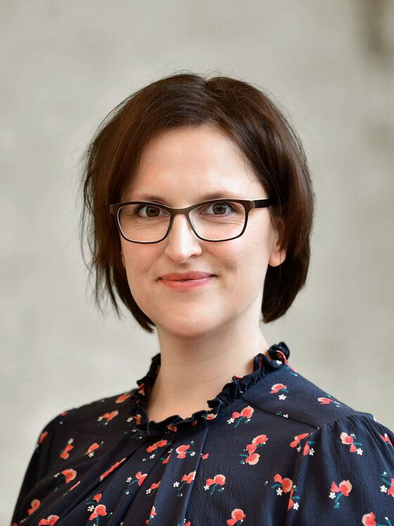 Eine weiblich gelesene Person mit kurzen braunen Haaren und Brille in blauer Bluse mit roten Applikationen lächelt vor grauem Hintergrund.