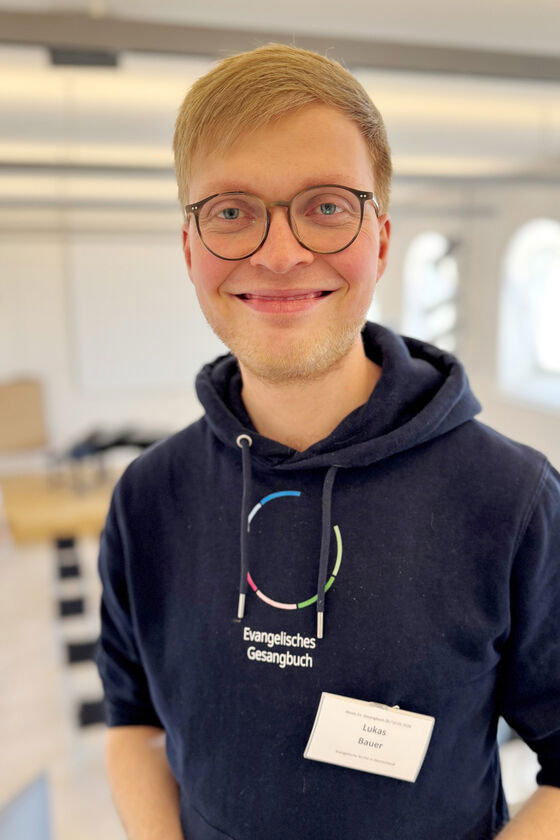 Portraitfoto einer jungen als Mann lesbaren Person mit kurzen Haaren und blauem Hoodie mit Gesangbuch-Logo