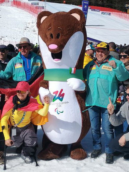 Mehrere Menschen stehen um das Maskottchen der Paralympics 2026 in Italien.