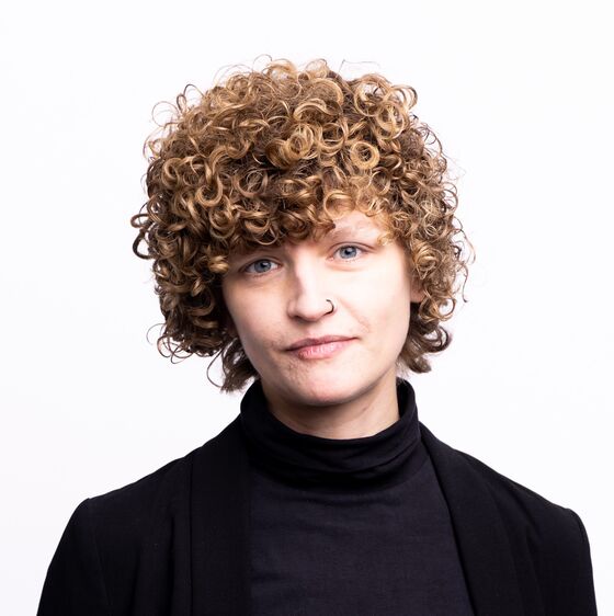 Das Bild zeigt eine weiblich gelesene Person mit Blick nach vorne. Sie trägt einen schwarzen Pullover. Der Hintergrund ist weiß. Die Haare sind lockig.