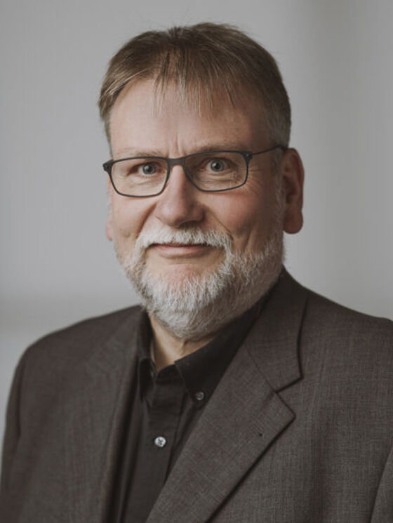 Auf dem Bild ist eine männlich gelesene Person. Sie trägt eine dunkle Brille, ein dunkles Hemd und ein dunkelgraues Sakko. Sie hat einen grauen Vollbart und kurze dunkle Haare. Der Schulter- und Kopfbereich ist vor einem grauen Hintergrund abgebildet.