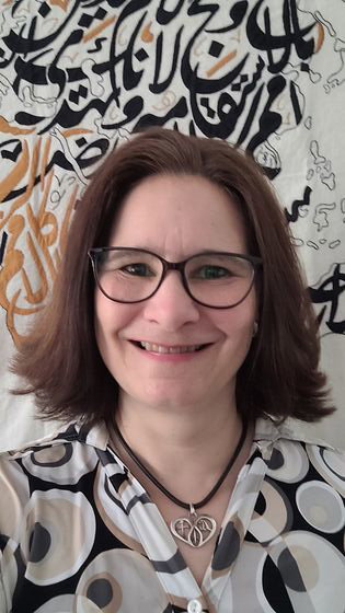 Auf dem Bild ist eine weiblich gelesene Person. Sie trägt eine Brille und hat schulterlanges, dunkles Haar. Sie steht vor einem abstrakt gestalteten Hintergrund in schwarz und beige.