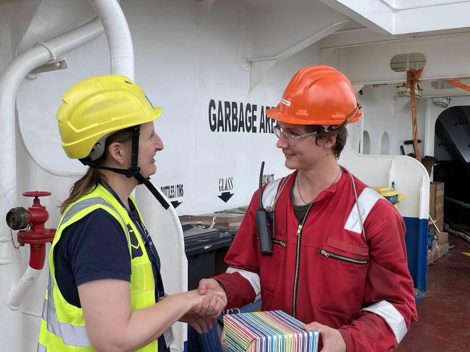 Eine weiblich gelesene Person, in gelber Schutzkleidung überreicht einer männlich gelesenen Person, in roter Schutzkleidung, ein Geschenk.