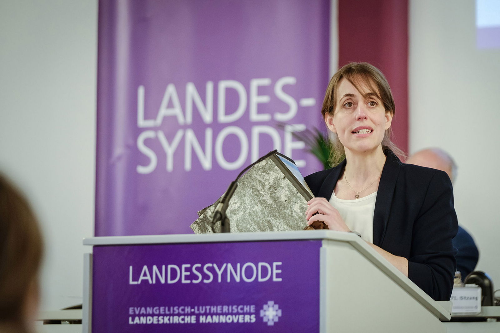 Eine weiblich gelesene Person an einem Redepult; sie hält einen Aktenordner in der Hand.