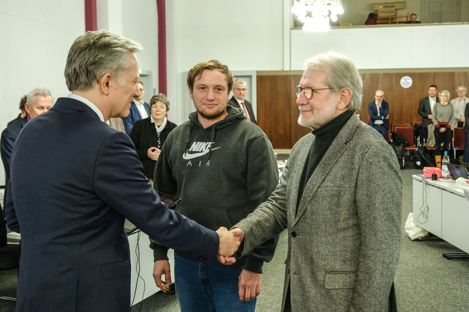 Drei männlich lesbare Personen stehen einander gegenüber, zwei geben sich die Hand. Sie stehen in einem Tagungsraum inmitten vieler Menschen. 