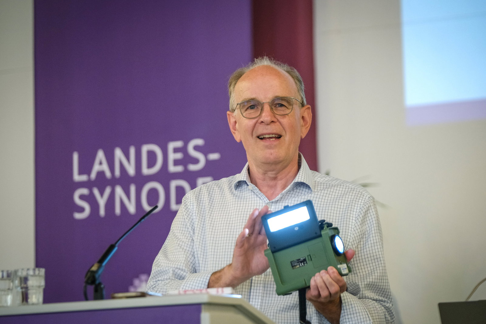 Eine männlich gelesene Person am Rednerpult mit einem Outdoor-Radio mit Solarzelle in der Hand.