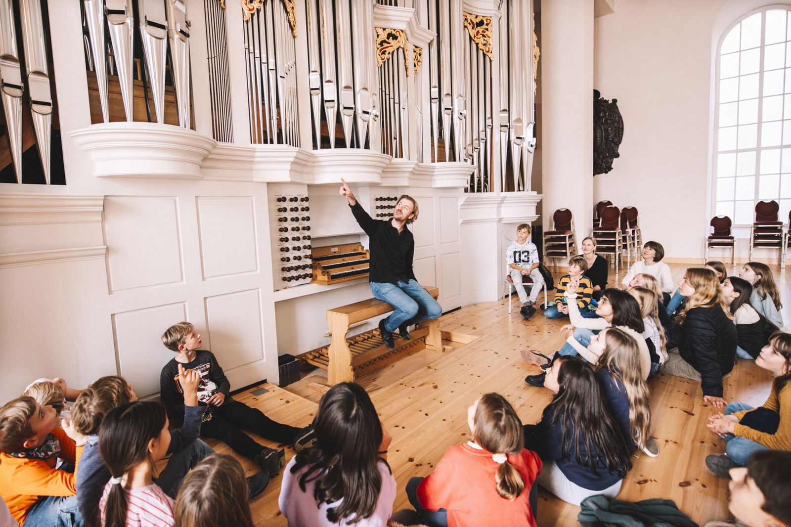 Eine Gruppe Kinder sitzt auf dem Boden vor einer Orgel. Auf der Orgelbank sitzt ein Mann, der nach oben deutet.