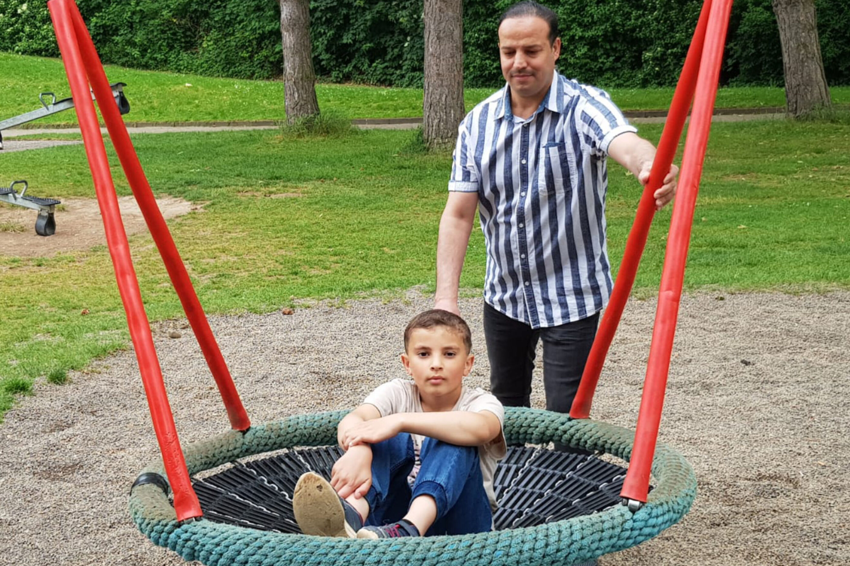 Vater und Sohn auf einem Spielplatz. Der Junge schaukelt, der Vater stößt ihn an.