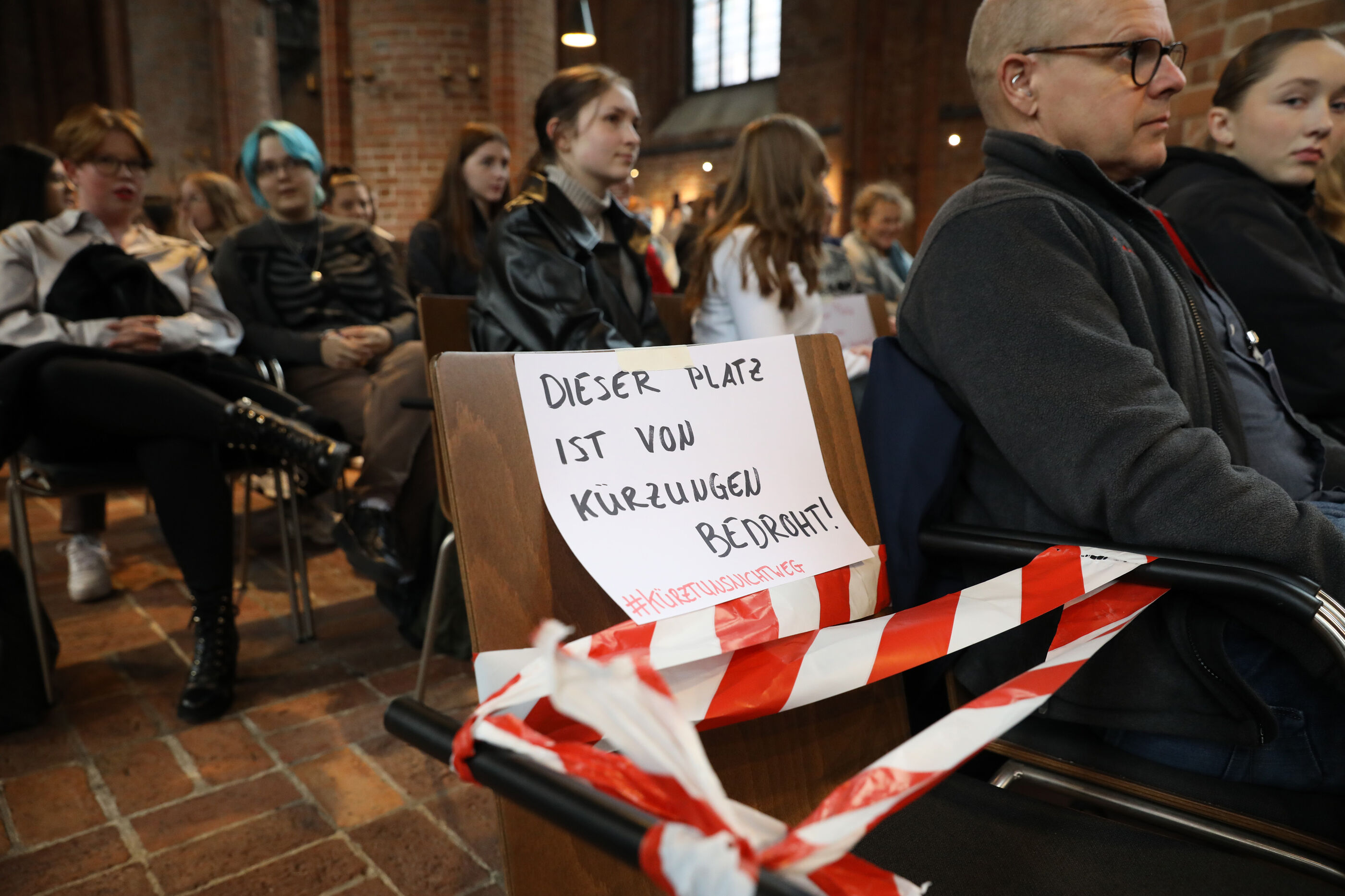 Ein Stuhl ist mit Flatterband abgesperrt. Darauf steht ein Schild mit der Aufschrift: Dieser Platz ist von Kürzungen bedroht.