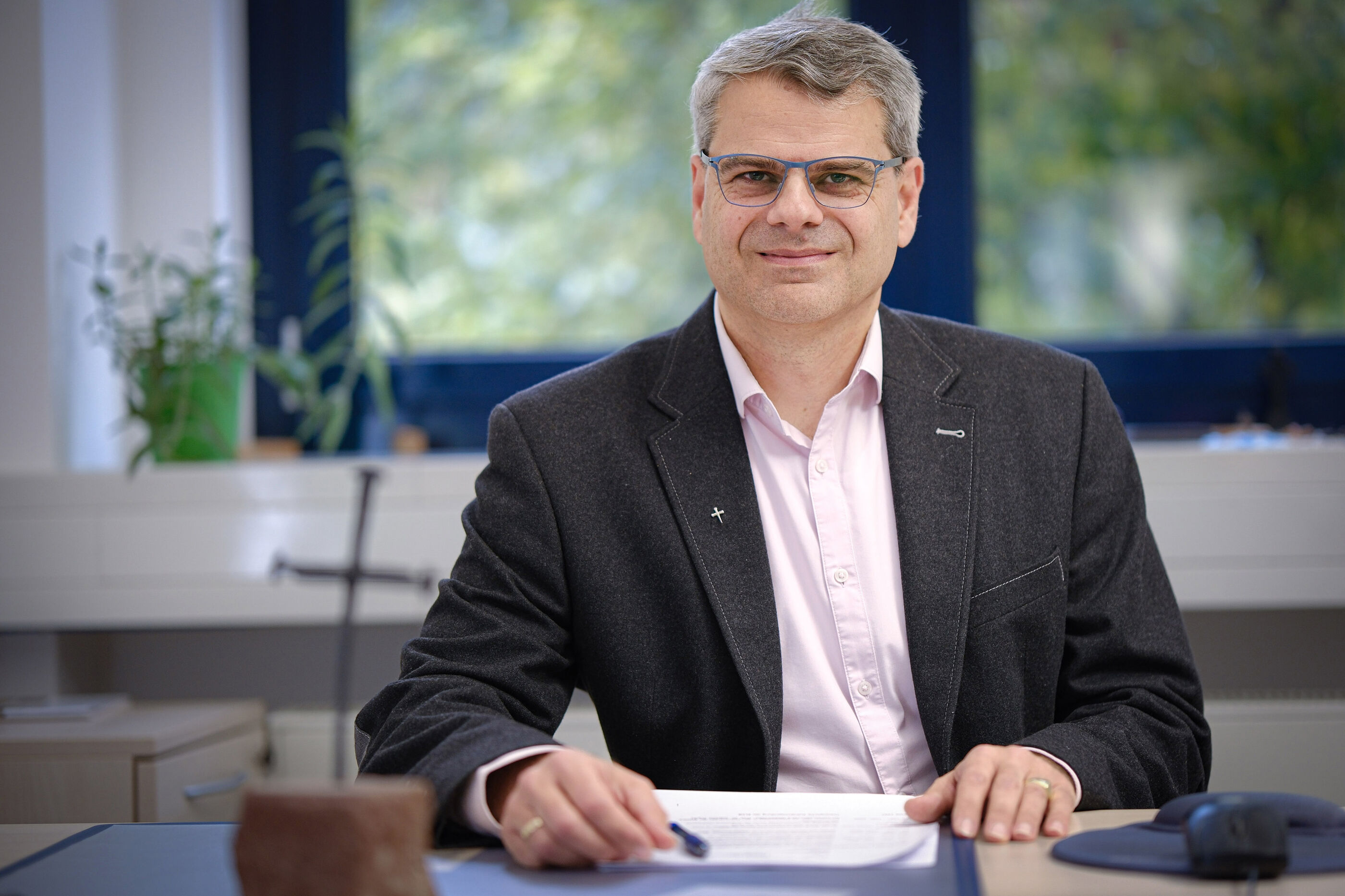 Ein mittelalter Mann mit kurzen Haaren, Brille und Sakko sitzt an seinem Schreibtisch vor Papieren.