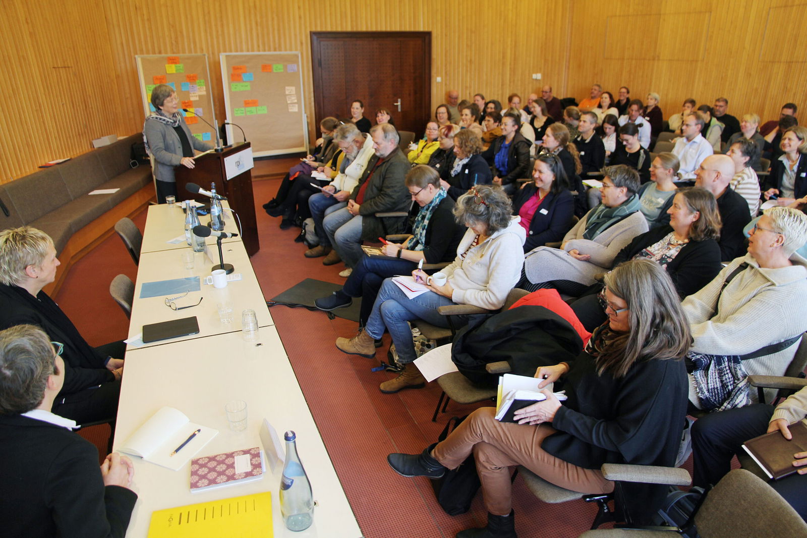 In einem Saal sitzen viele Menschen. Vorn sitzt die Tagungsleitung, am Redepult steht eine ältere Frau.