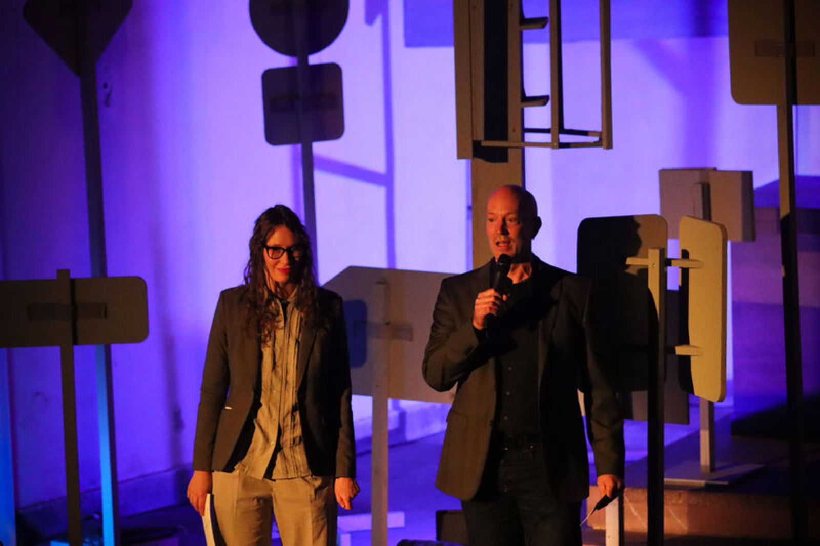 Eine Frau und ein Mann stehen auf einer Bühne im Dämmerlicht. Der Mann spricht in ein Mikrofon.