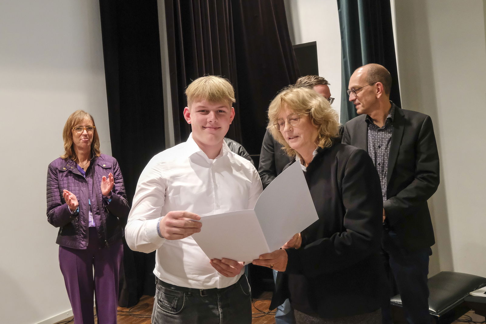 Eine blonde Frau übergibt einem jungen Mann in weißem Hemd eine weiße Urkundenmappe. Im Hintergrund stehen eine Frau in lila Anzug und ein Mann in dunklem Sakko.
